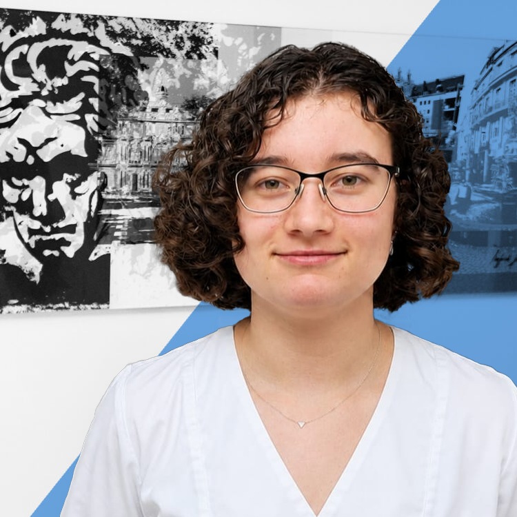 Portrait of a young person with short curly brown hair, glasses, and a light blue blouse, smiling softly in front of a graphic mural with blue shapes.